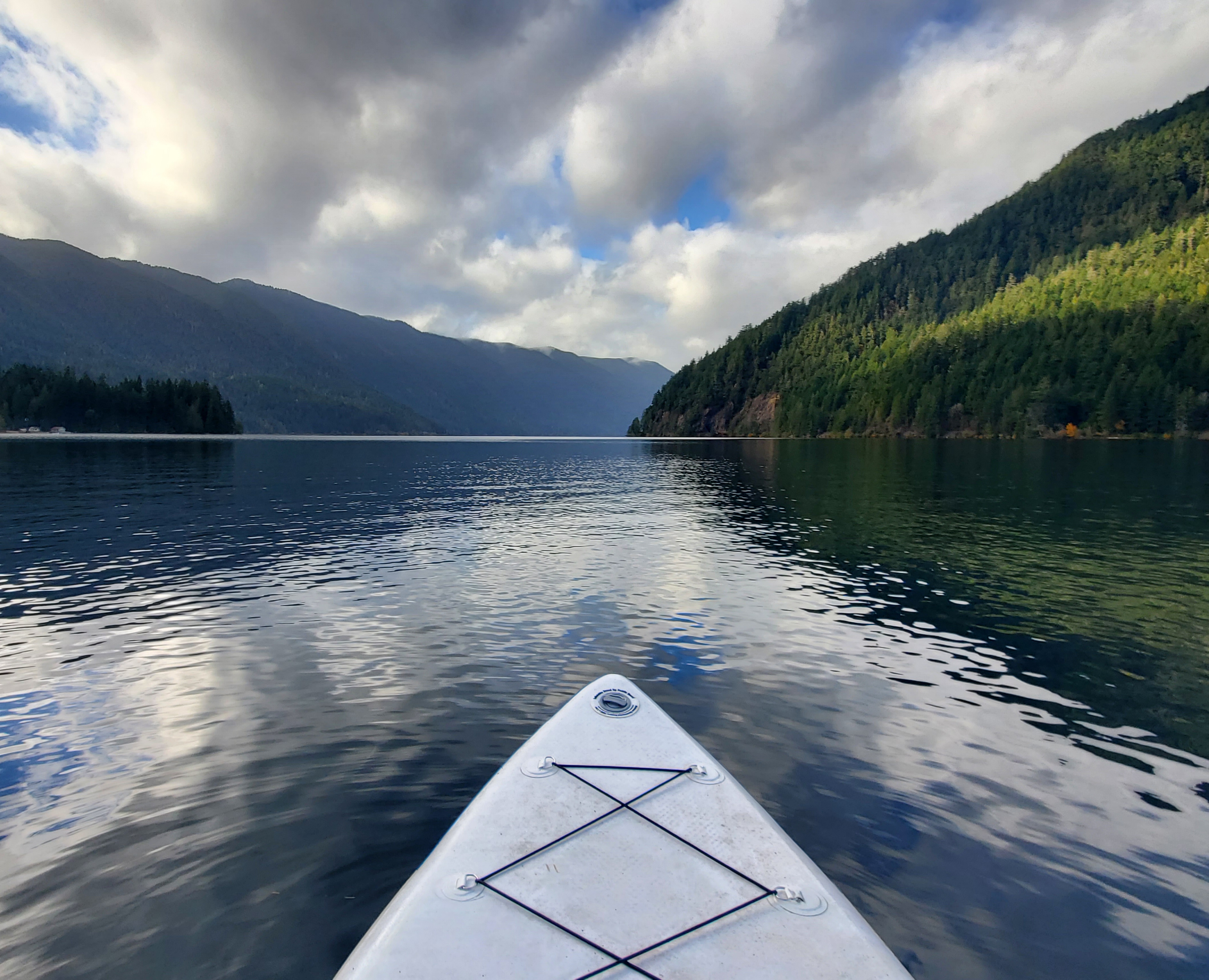 Photograph of the end of an inflatable paddleboard pointed toward a lake view, surrounding green mountains and a partly cloudy sky.
